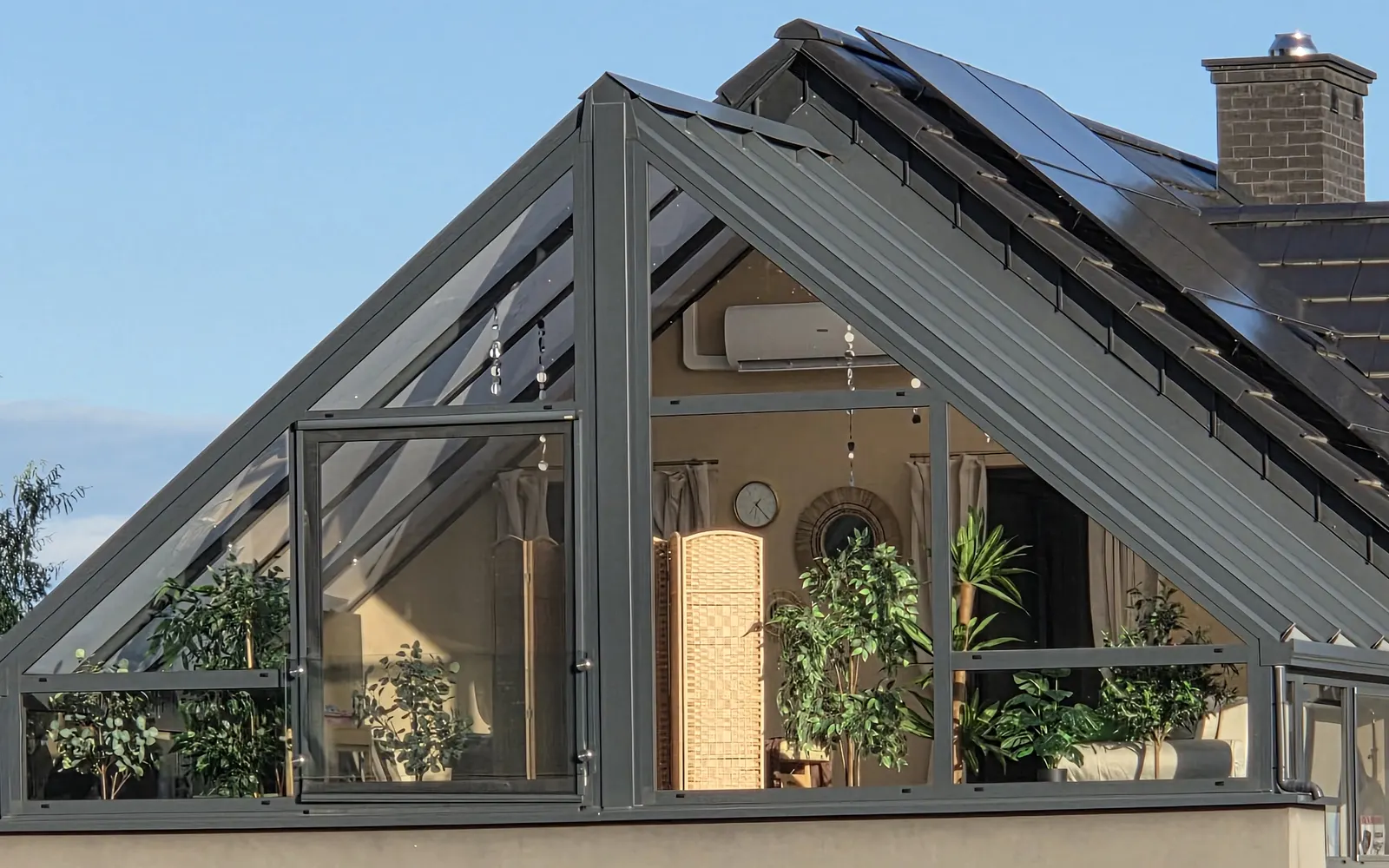 A glass greenhouse on the upper floor of a house, with green plants, a folding screen and soft light inside